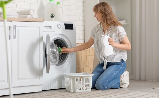 Vrouw knielt voor een wasmachine en stopt wasgoed in de trommel terwijl ze een fles wasmiddel vasthoudt