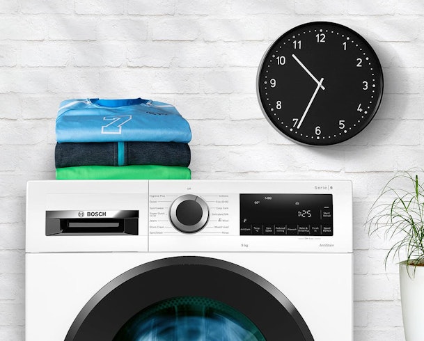 Bosch wasmachine met SpeedPerfect functie onder wandklok en opgevouwen wasgoed