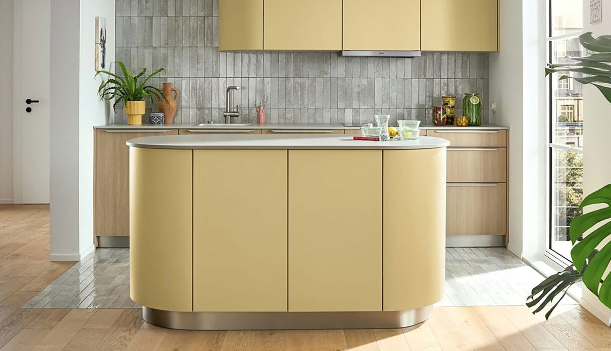 Pastelgele keuken met afgerond kookeiland, licht houten kasten en grijze tegelwand.
