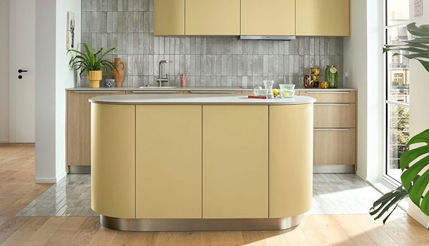 Pastelgele keuken met afgerond kookeiland, licht houten kasten en grijze tegelwand.