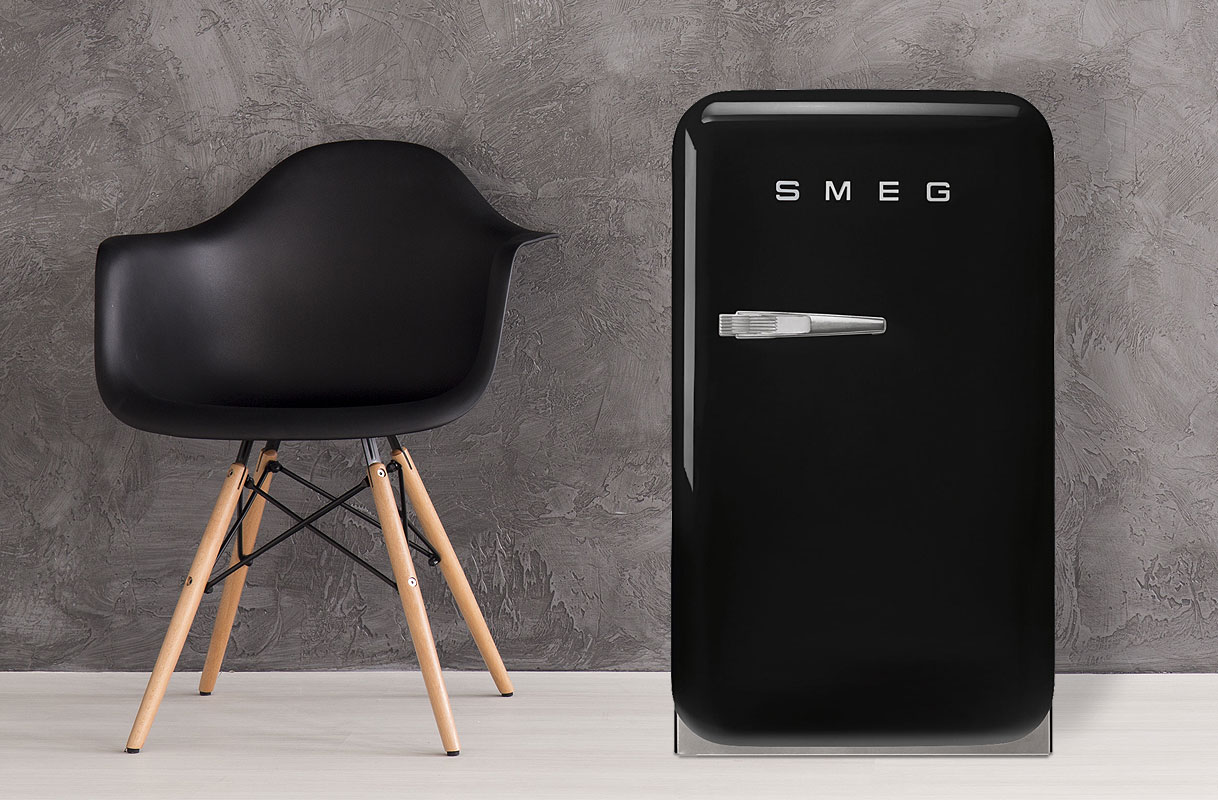 Zwarte tafelmodel koelkast in retrodesign van Smeg naast moderne zwarte stoel met houten poten tegen grijze betonnen muur.